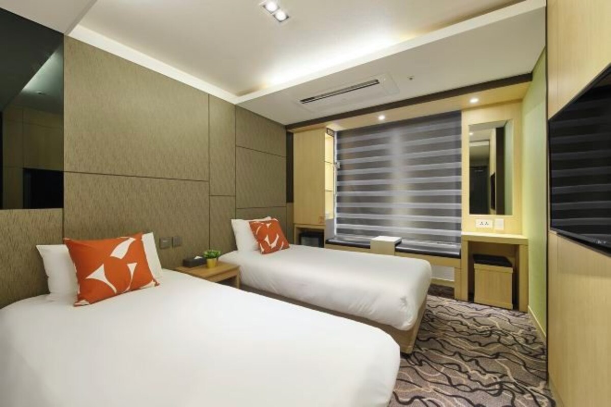 A hotel room with two beds, a small desk, a mirror, and a window with blinds.