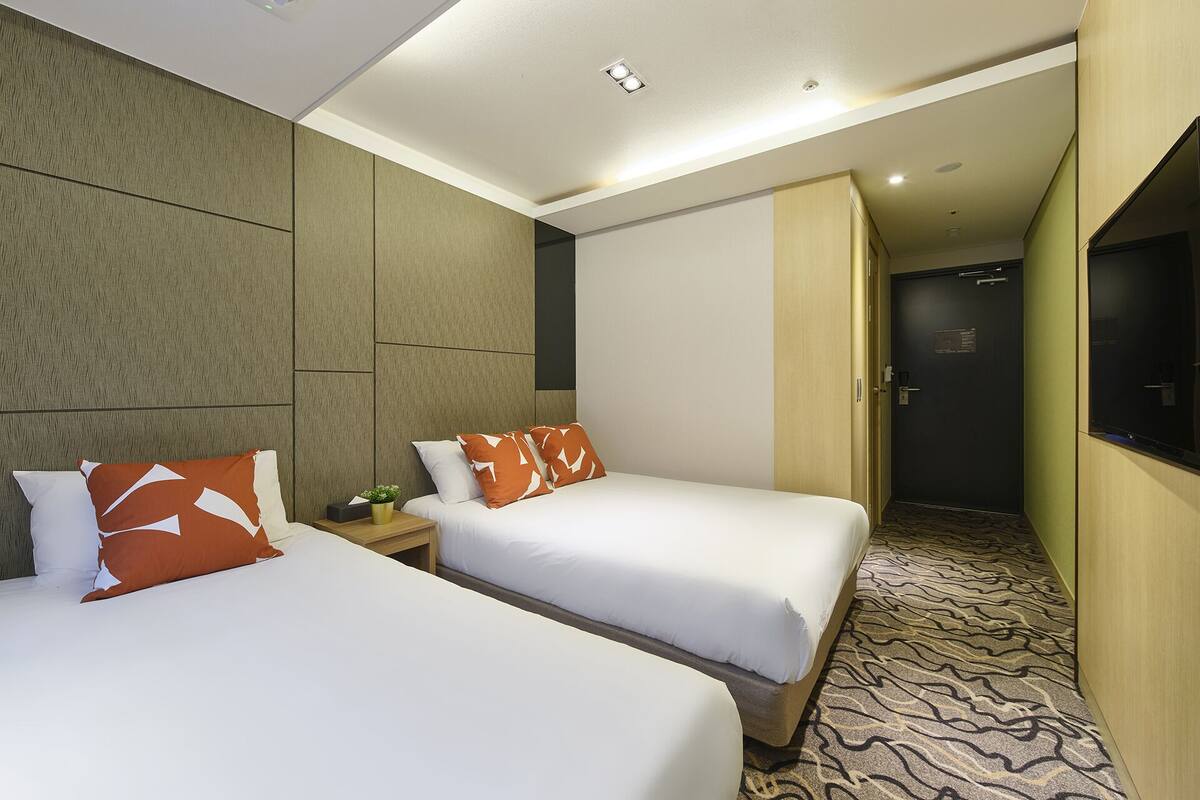 Habitación de hotel con dos camas, una mesita de noche con lámpara, un televisor montado en la pared y una puerta que da a otra habitación.