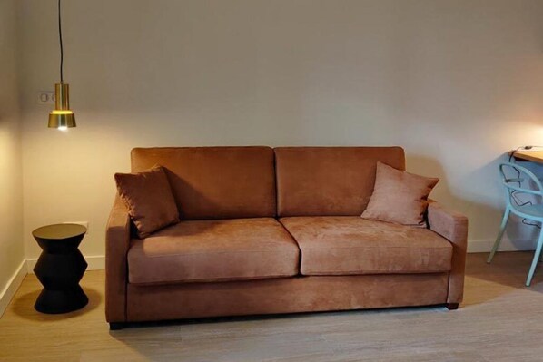 A brown sofa with two cushions, a small black side table, and a hanging light fixture. - Studio SUP N#2 (Narbonne)