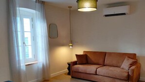 A living room with a brown sofa, a small side table, a lamp, and a window with sheer curtains. - Double Appartement #1 (Narbonne)