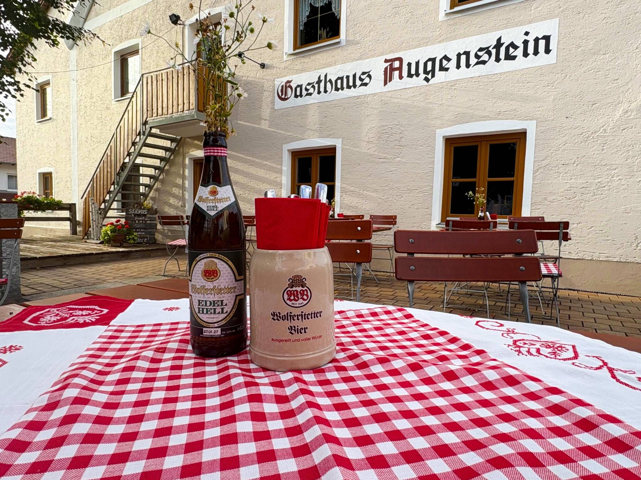 Ein Tisch mit karierter Tischdecke, zwei Bierflaschen und ein Schild mit der Aufschrift 'Gasthaus Augenstein' an einem Gebäude.