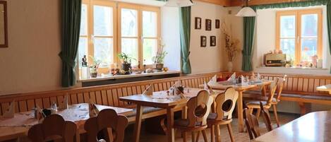 A cozy restaurant with wooden furniture, large windows with curtains, and a variety of plants on the windowsill.