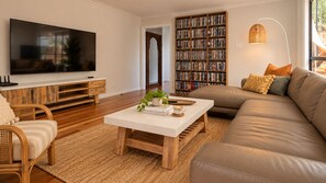 A living room with a flat-screen TV, a bookshelf, a sofa, and a coffee table. - Splendour Hideaway - Luxury and pool - Caves Beach (Caves Beach)