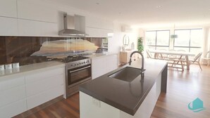 A modern kitchen with white cabinetry, a central island, and a large window providing natural light. - Splendour Hideaway - Luxury and pool - Caves Beach (Caves Beach)