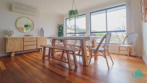 A dining area with a wooden table and chairs, a white cabinet, and large windows offering a view of the outdoors. - Splendour Hideaway - Luxury and pool - Caves Beach (Caves Beach)