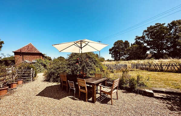 A rustic outdoor dining area with a wooden table and chairs, a large umbrella, and a brick building in the background.