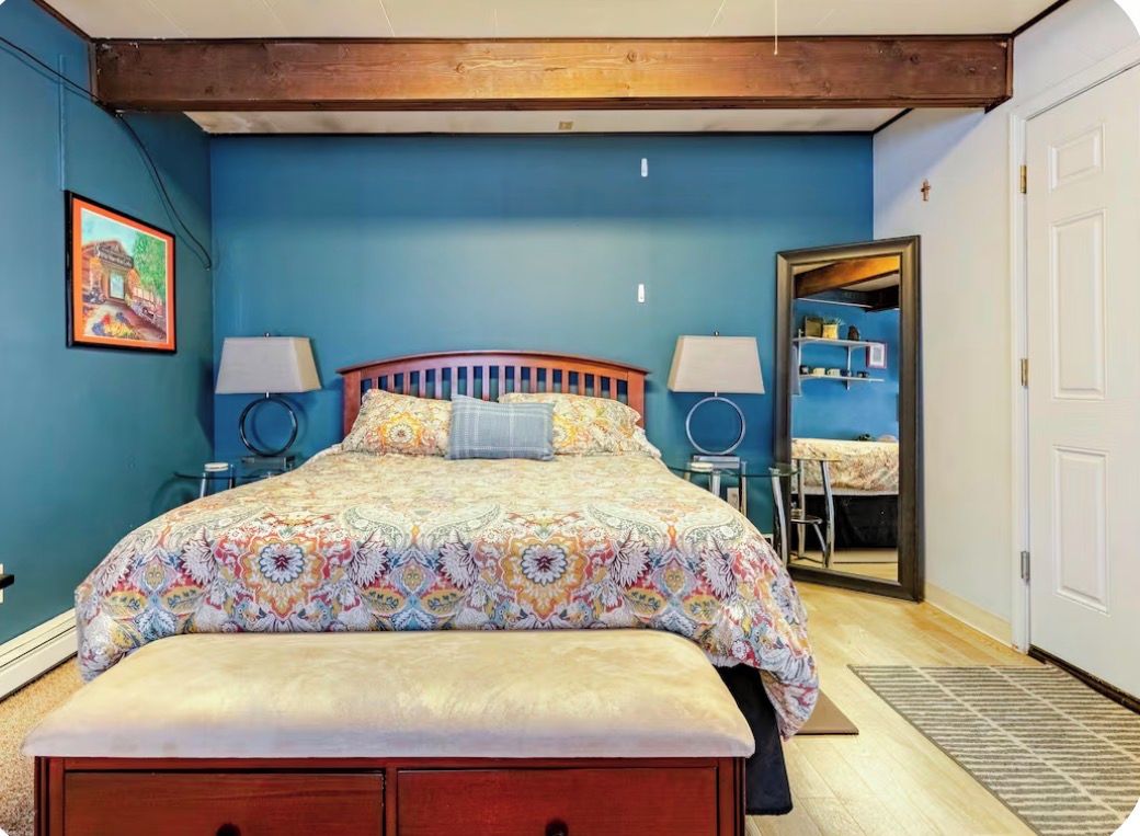 Un dormitorio con cama, dos lámparas de noche, cabecera de madera, un banco y una puerta que da al baño.