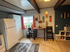 Una cocina compacta con refrigerador, una mesa de comedor con sillas, un ventanal con cortinas y una pared decorada con diversos adornos.