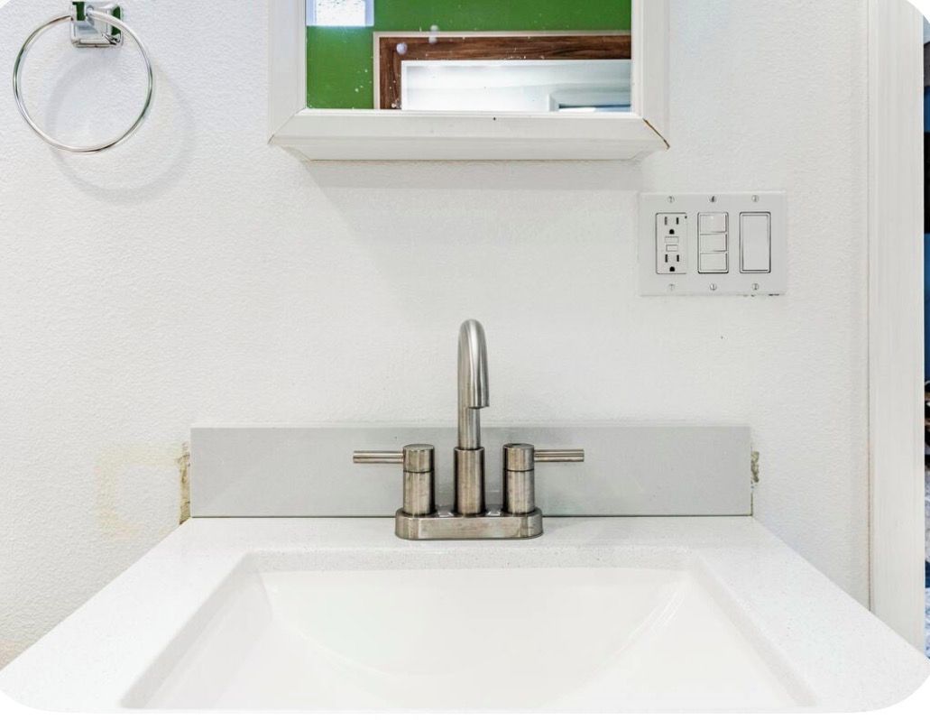 Un bagno con un lavandino bianco, un rubinetto, una finestra sopra il lavandino e una presa di corrente sulla parete.