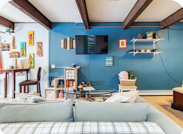 A room with a blue wall, a flat-screen TV mounted above a shelf, a sofa with patterned cushions, and a wooden ceiling.
