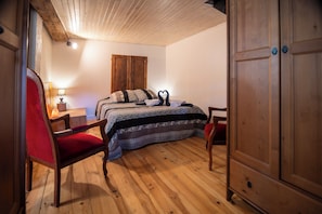 Une chambre avec un plancher en bois, un lit avec une literie à motifs, une chaise rouge, une armoire en bois et une table de chevet avec une lampe.