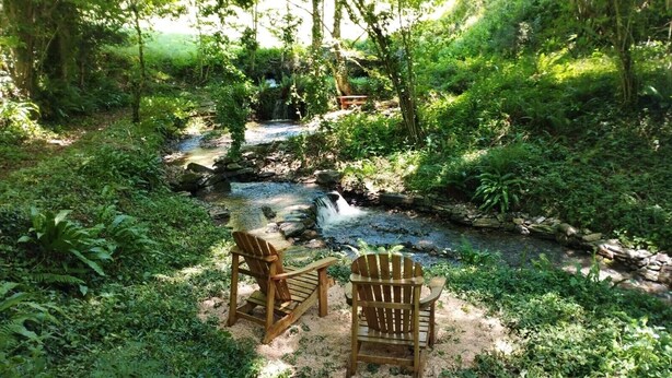 Eine friedliche Umgebung im Freien mit einem kleinen Wasserfall, zwei hölzernen Adirondack-Stühlen und einem Kiesweg.