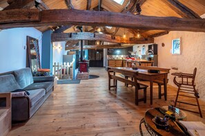 Un intérieur rustique avec un plafond en bois, un coin repas avec une table et des bancs en bois, une cuisine équipée d’appareils modernes et un coin salon confortable avec un canapé et des coussins.