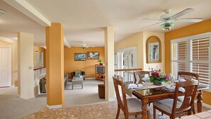 A dining area with a wooden table and chairs, a ceiling fan, and a view of the living room in the background.