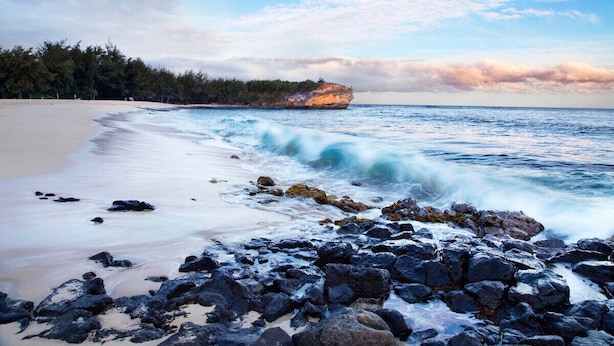 A beach with a rocky foreground, a sandy shore, and a large rock formation in the distance.