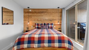 A bedroom with a wooden headboard, a checkered bedspread, two pendant lights, and a large window with a view.