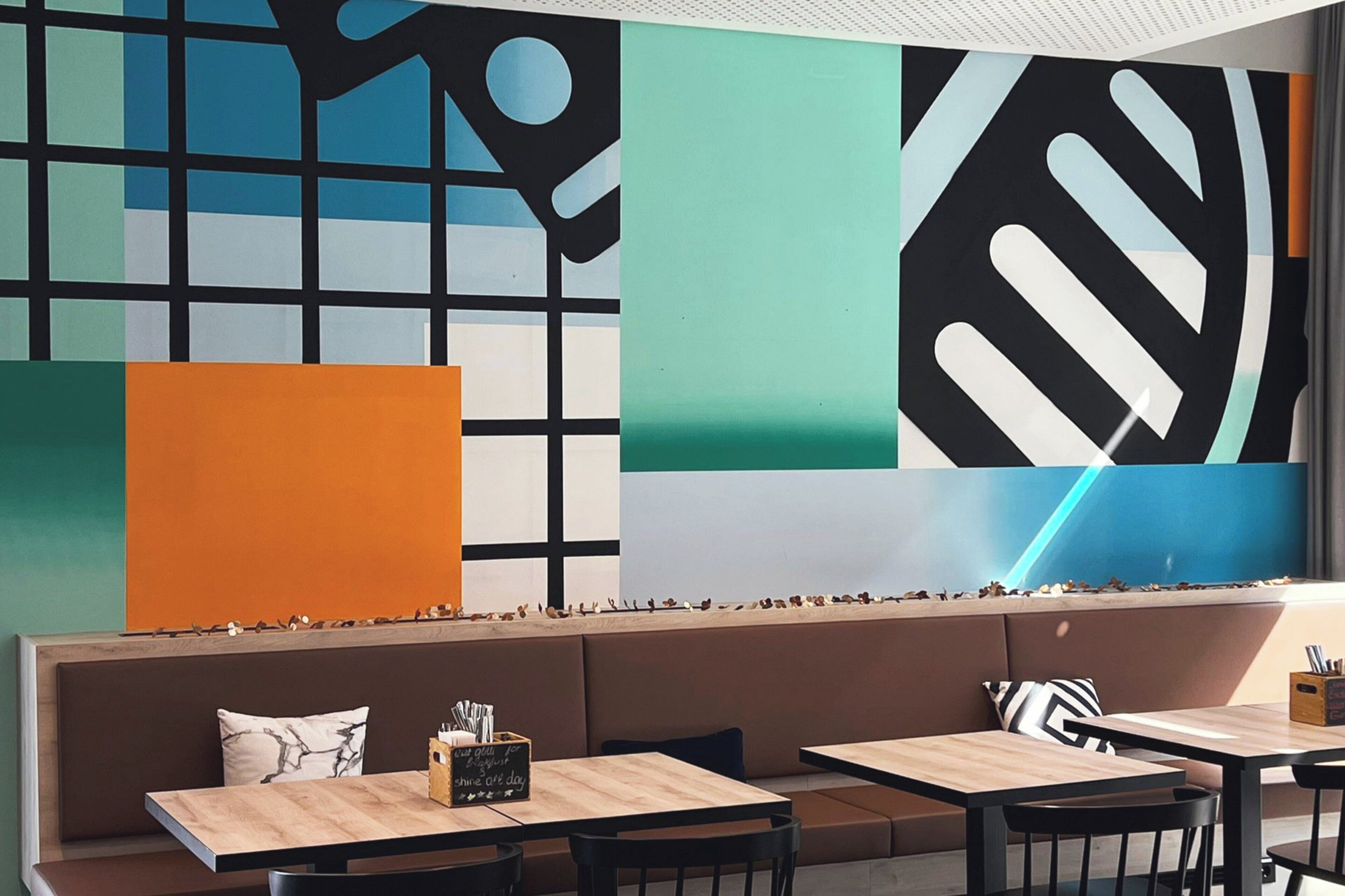A modern interior with a long bench seating area, wooden tables, and a large abstract wall mural.