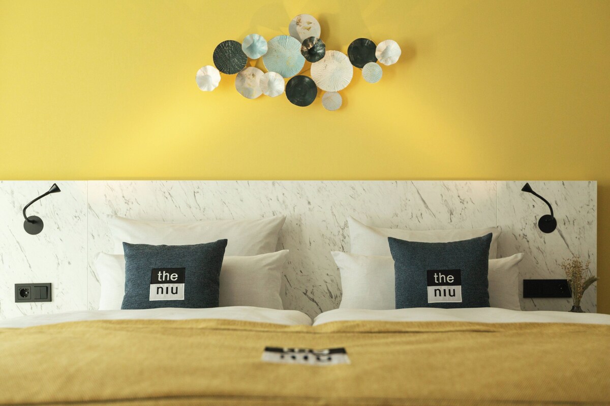 Un llit amb dos almoinos, una làmpada penjada a la paret i una obra d'art decorativa a la pared.