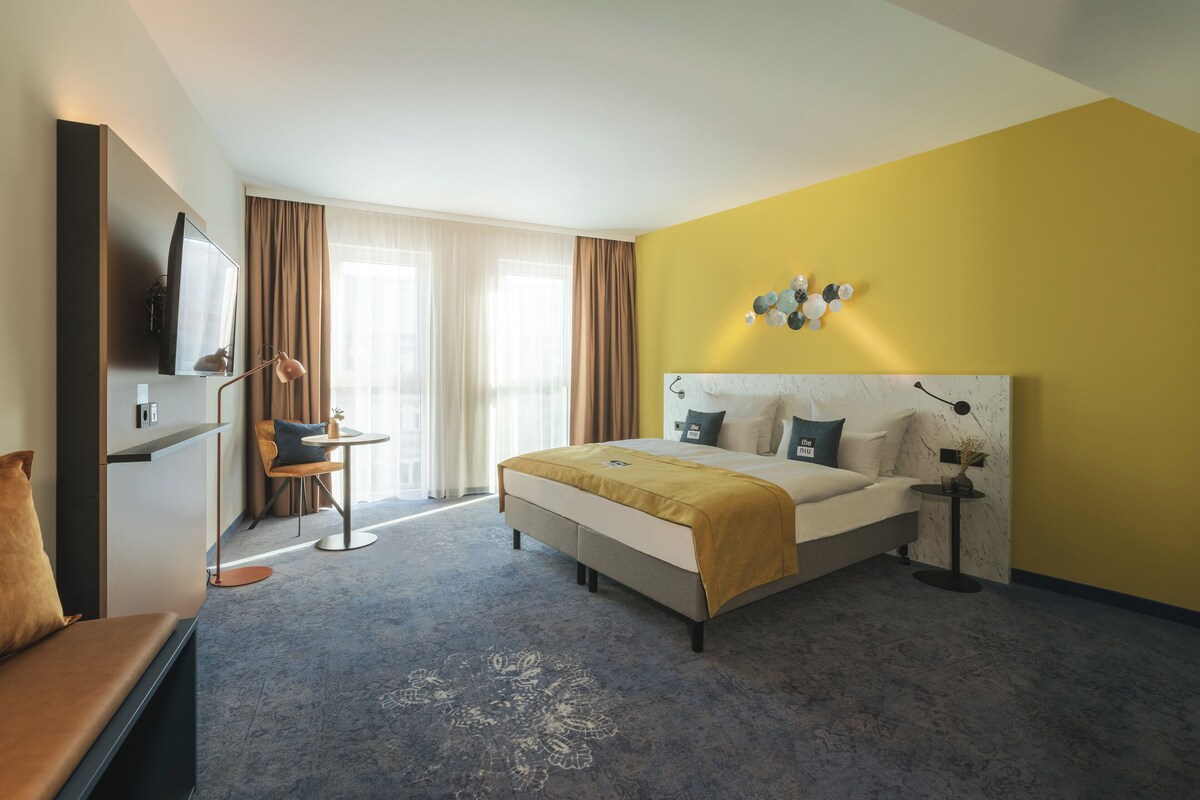 Habitación de hotel con cama, escritorio, silla, televisor y ventana con cortinas.