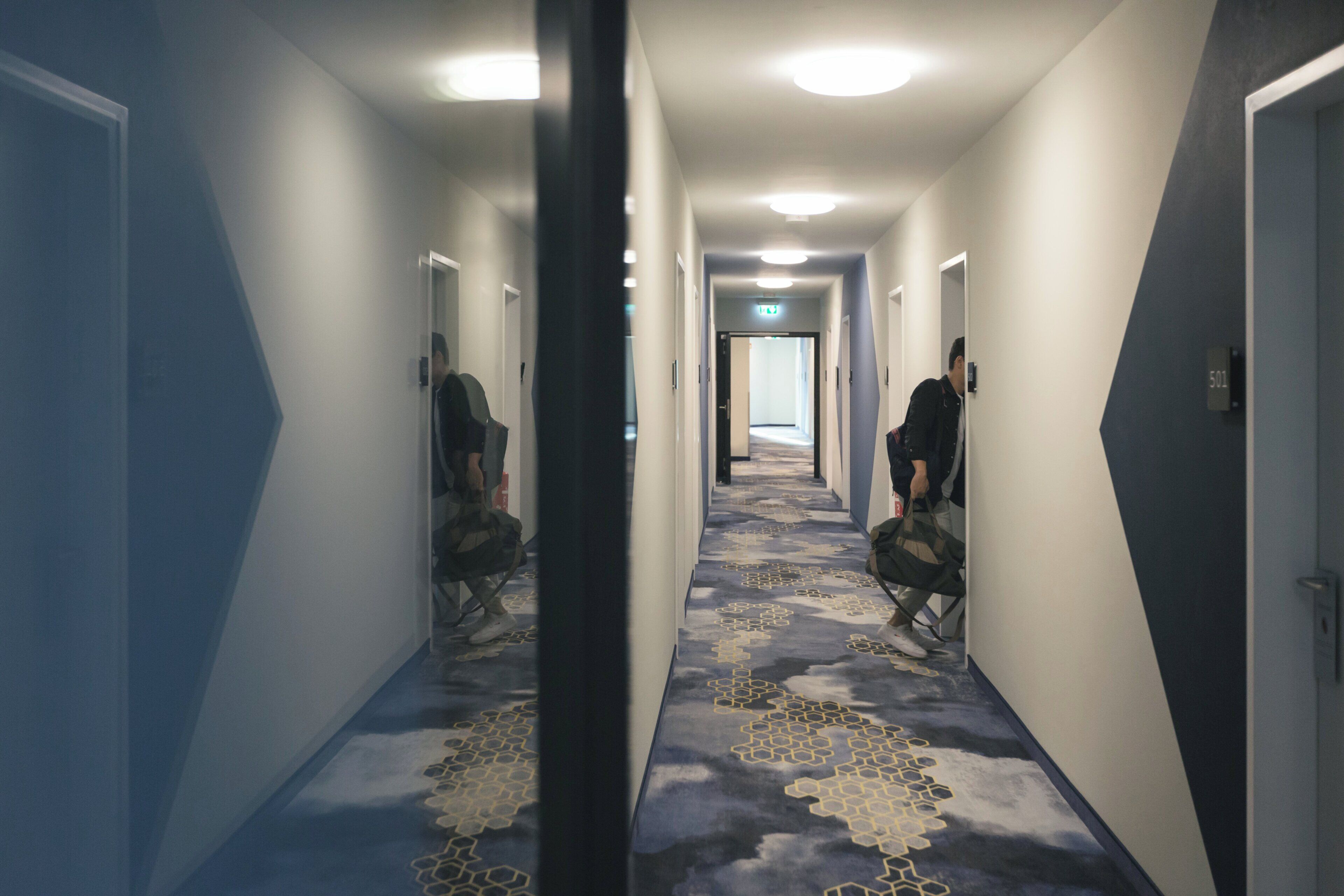 Un couloir avec un tapis à motifs, des portes de chaque côté et une personne assise sur une chaise.