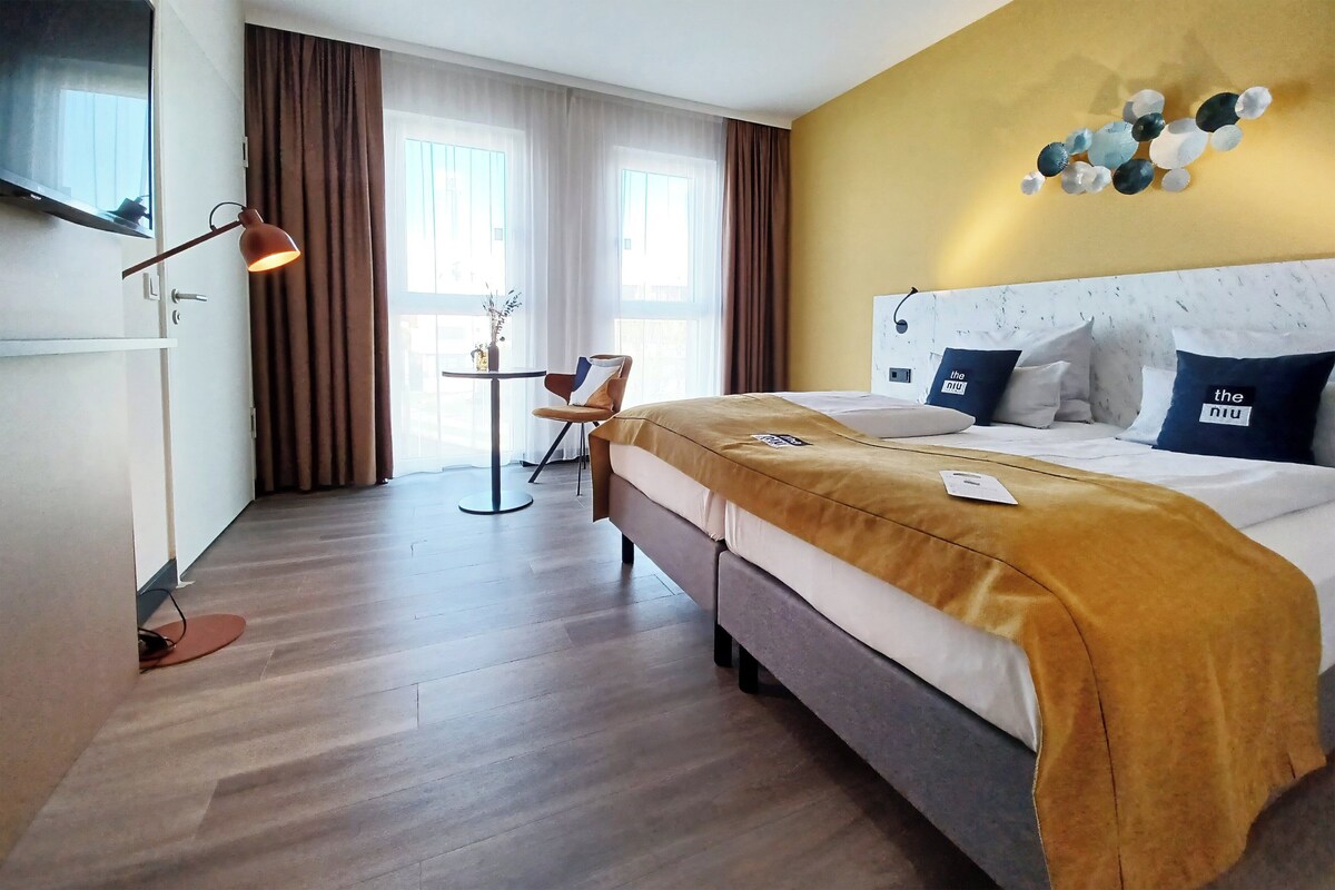 Una habitación de hotel con cama, escritorio, silla, televisor y ventana con cortinas.