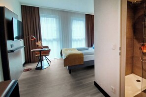 Habitación de hotel con cama, escritorio, silla, televisor y zona de ducha.