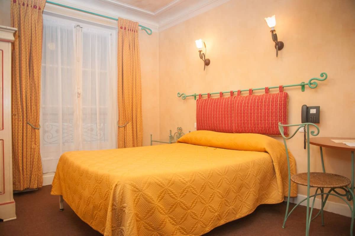 A room with a bed, a table, and a chair. There are curtains on the windows and a wall-mounted telephone.
