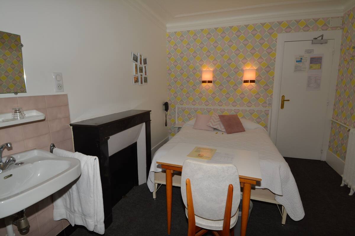 A hotel room with a bed, a bedside table, a chair, a sink, and a mirror.