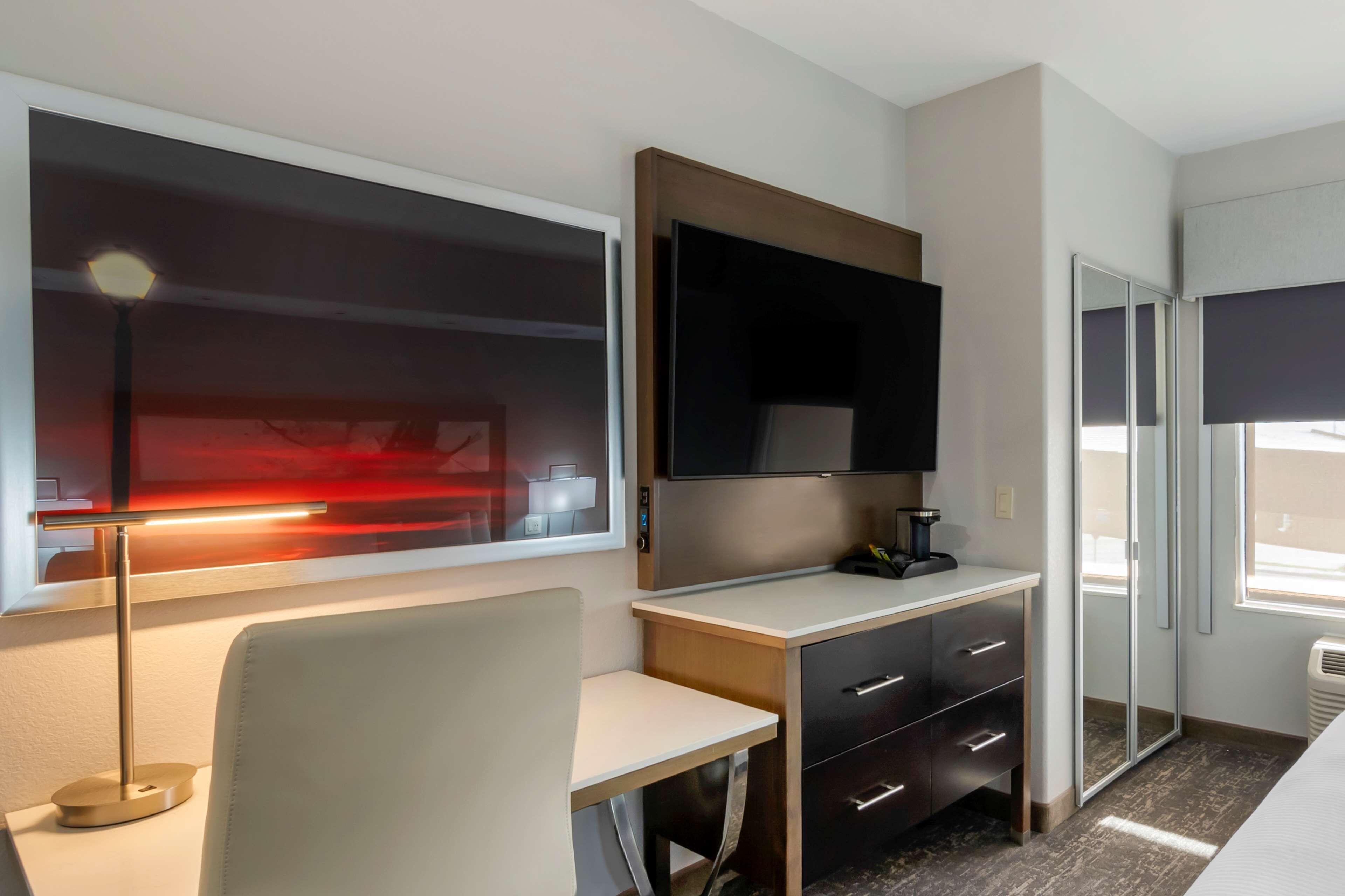 A modern hotel room with a desk, a flat-screen TV, a bedside table, and a wardrobe.