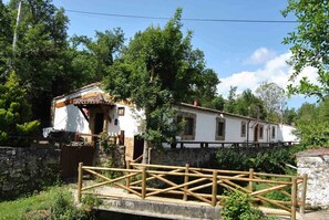 Un edificio rústico con fachada blanca y tejado de tejas, rodeado de exuberante vegetación y un muro de piedra.