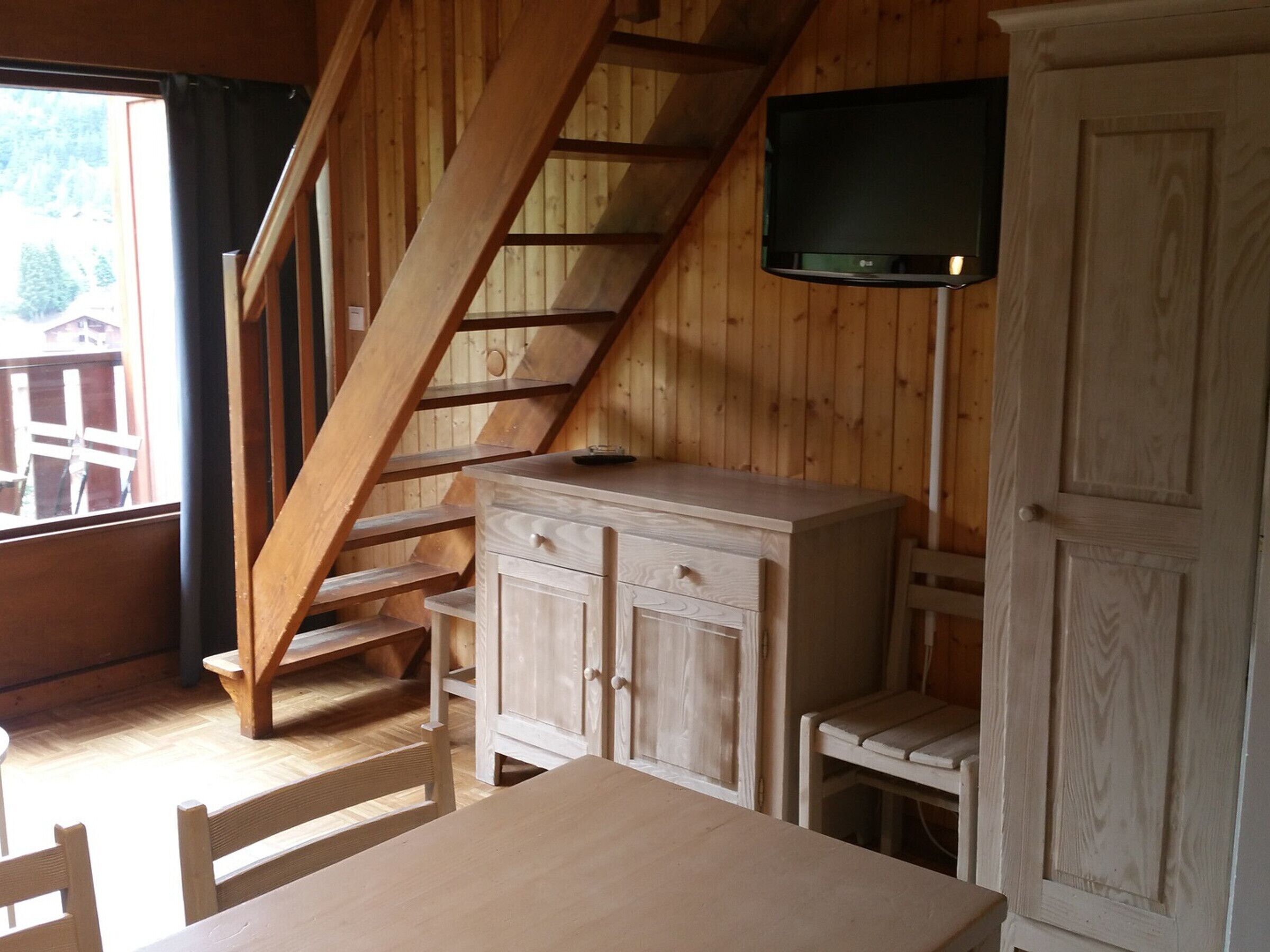 A wooden interior with a staircase, a cabinet, a television mounted on the wall, and a window with a view of trees.