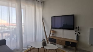 Moderni olohuone, jossa on sohva, seinälle asennettu televisio, pieni pöytä lampun kanssa ja näkymä ulos läpikuultavien verhojen kautta.