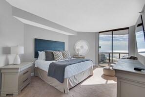 Une chambre avec un grand lit, une table de chevet, une lampe, un élément de décoration circulaire fixé au mur et une vue sur la plage, visible à travers une grande fenêtre.