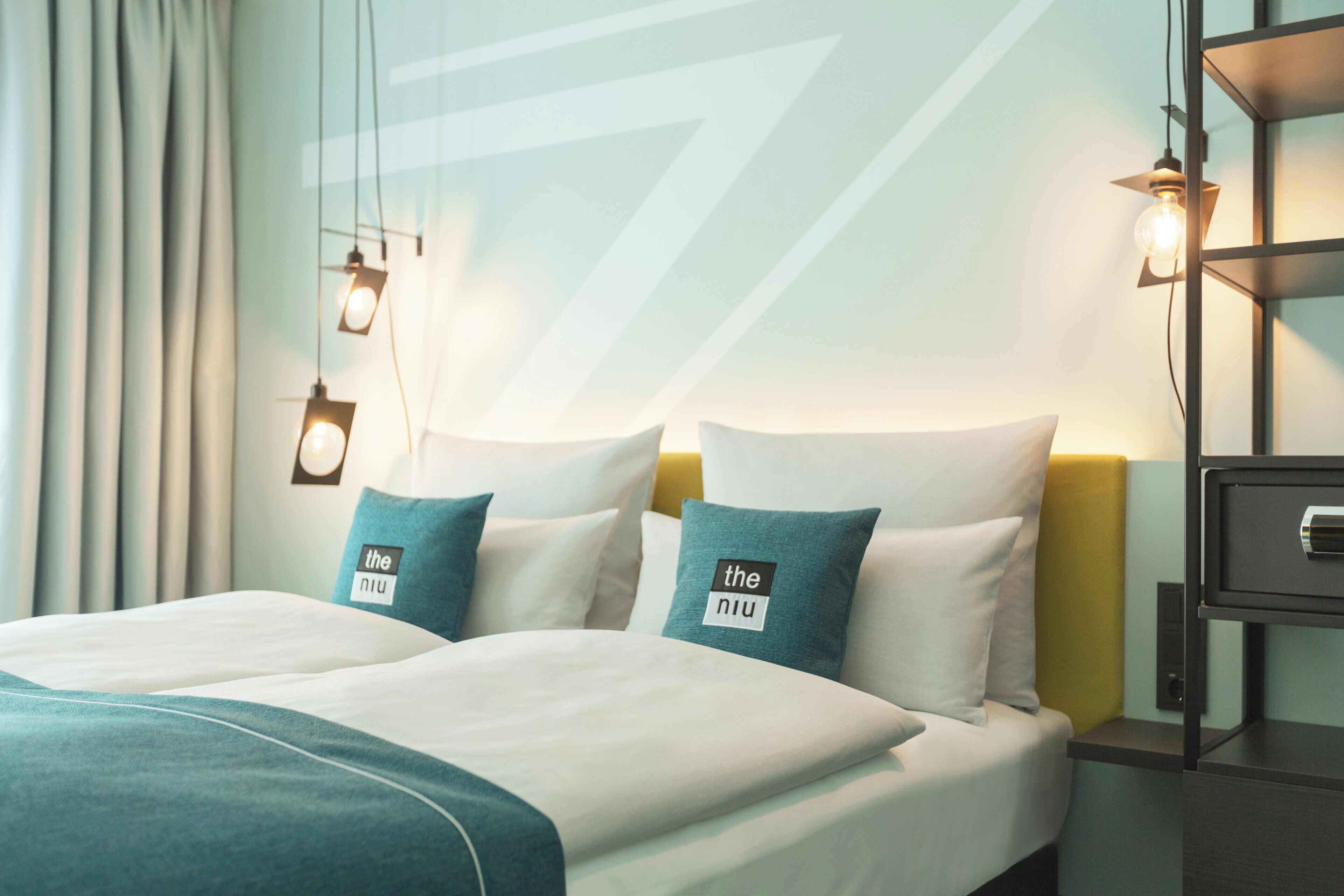 A hotel room with a bed, two pillows with a logo, a nightstand, and a wall-mounted lamp.