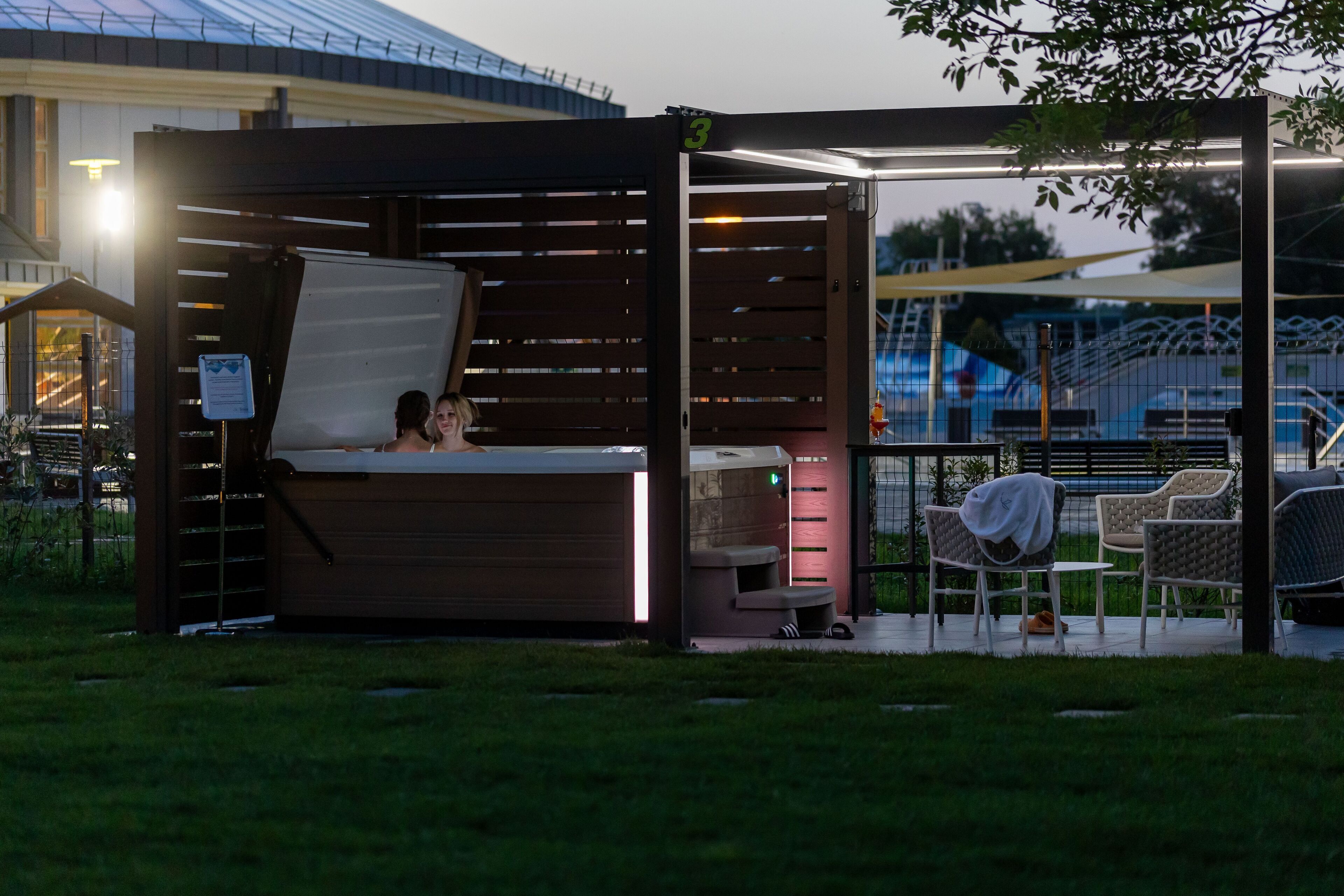 A small, enclosed outdoor seating area with a bench and chairs, a covered table, and a view of a sports field.