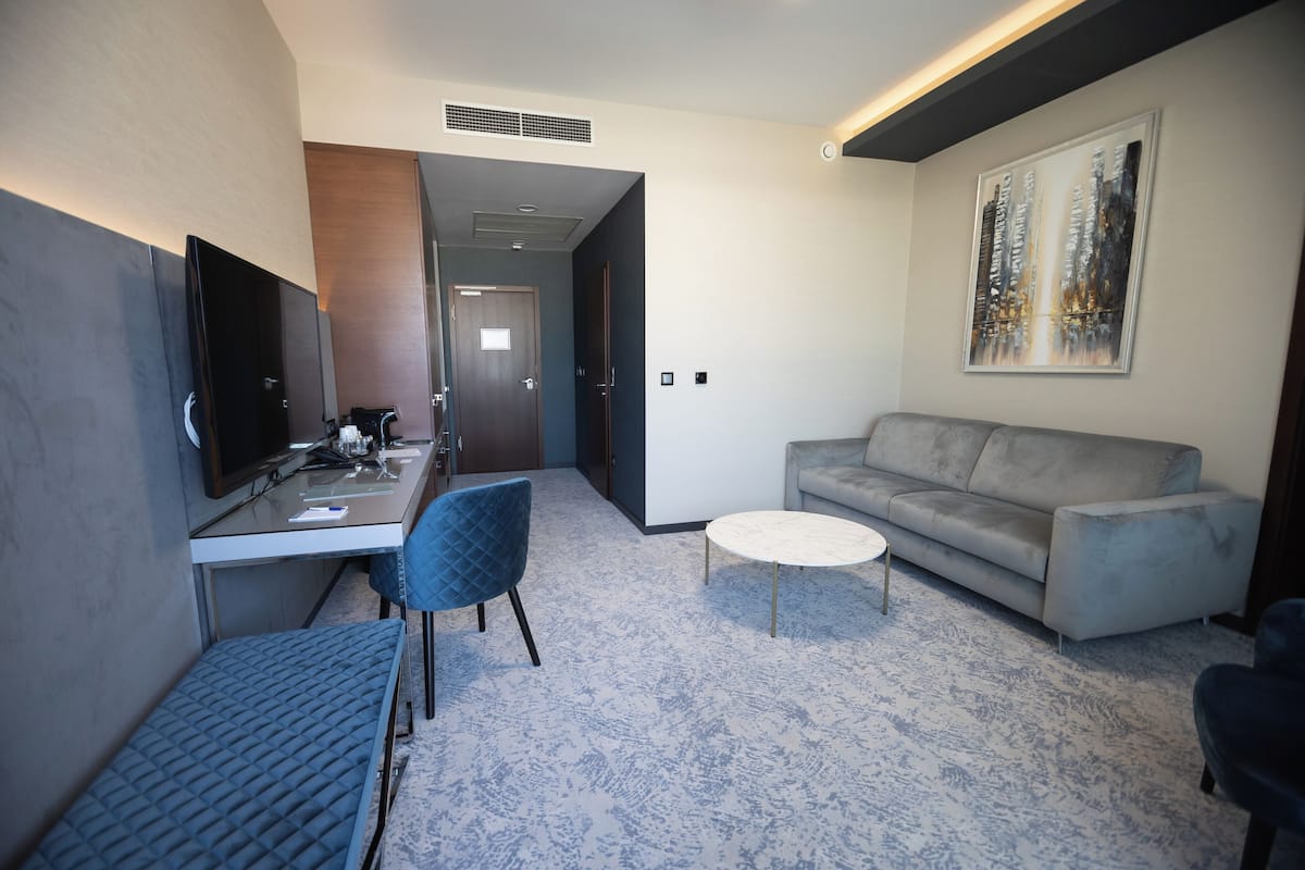 Una habitación de hotel moderna con televisión de pantalla plana, un escritorio, una silla con cojín azul, un sofá gris y una mesa de centro redonda.