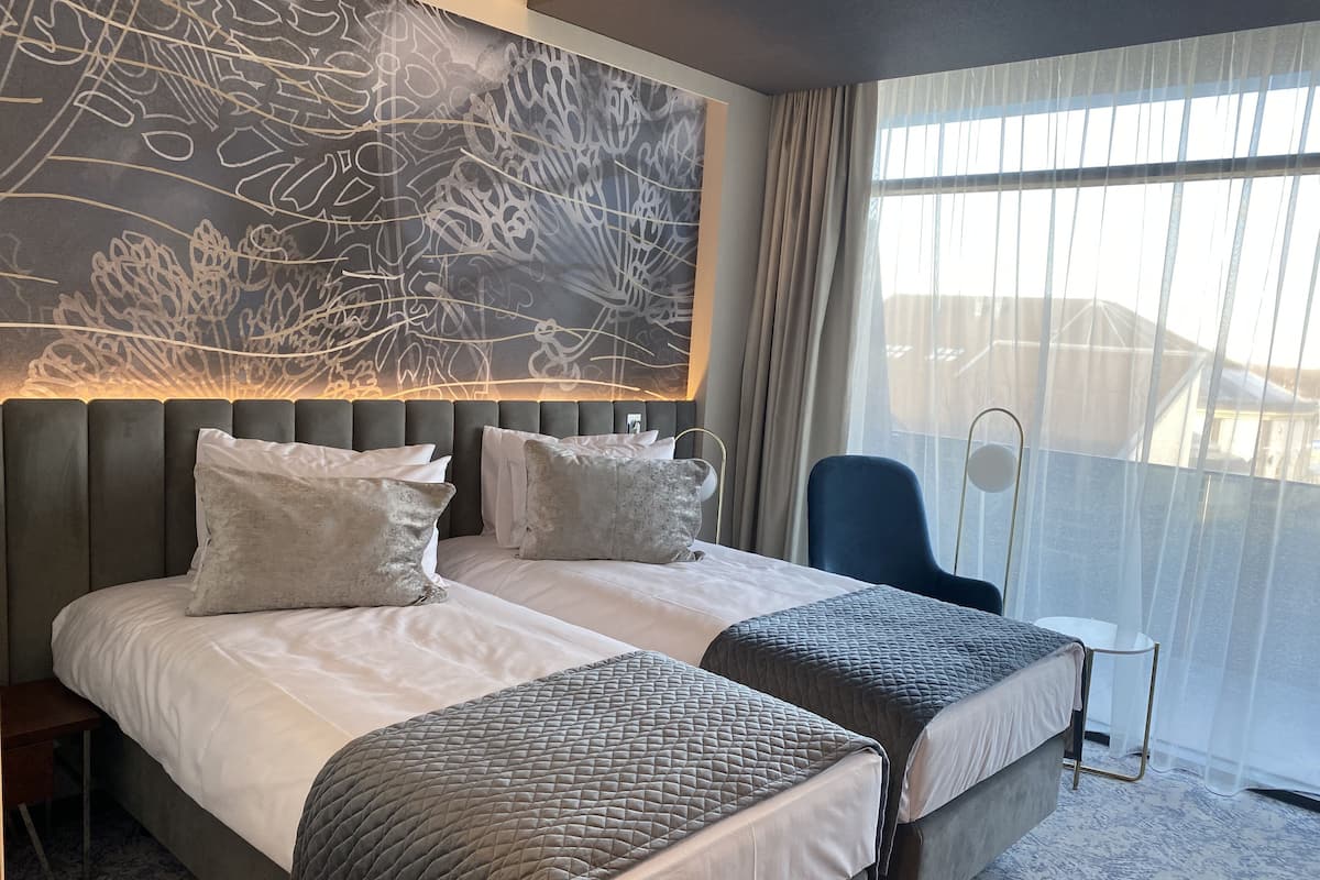 Habitación de hotel con una cama grande, dos mesitas de noche, una silla y un ventanal con cortinas.