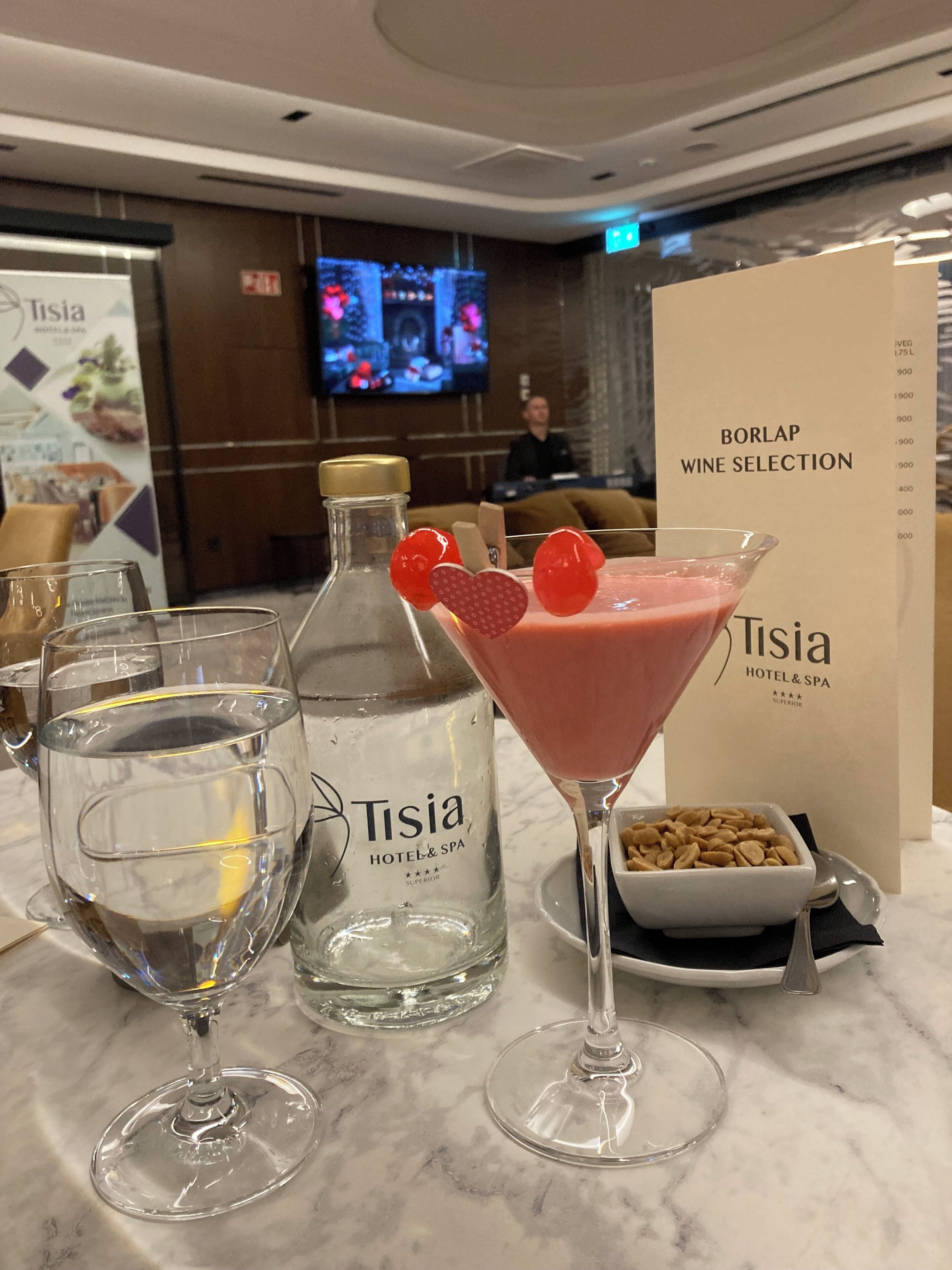 Una zona de bar con un menú de vinos Borlap, un cartel del Hotel & Spa Tisia, una botella de Tisia y una copa de martini con adornos.