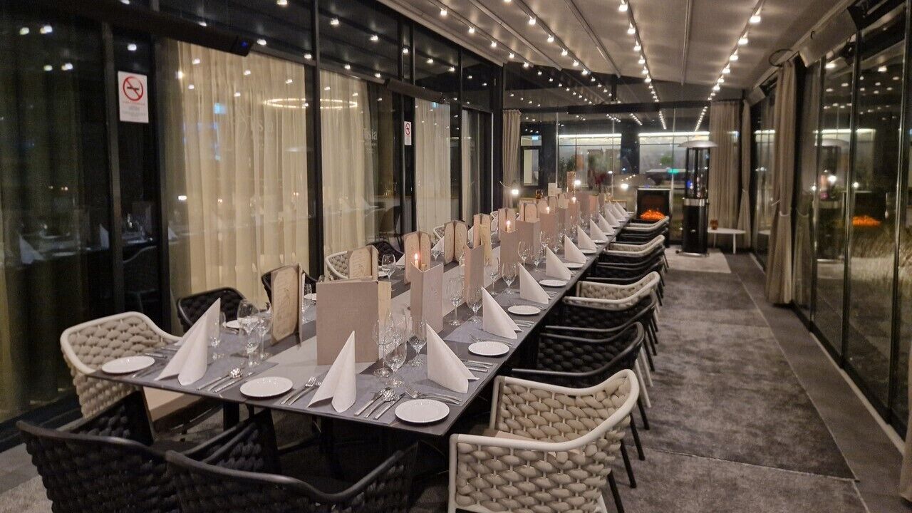 A long dining table set with white napkins, plates, and cutlery, flanked by wicker chairs, in a modern indoor setting with glass partitions and a carpeted floor.
