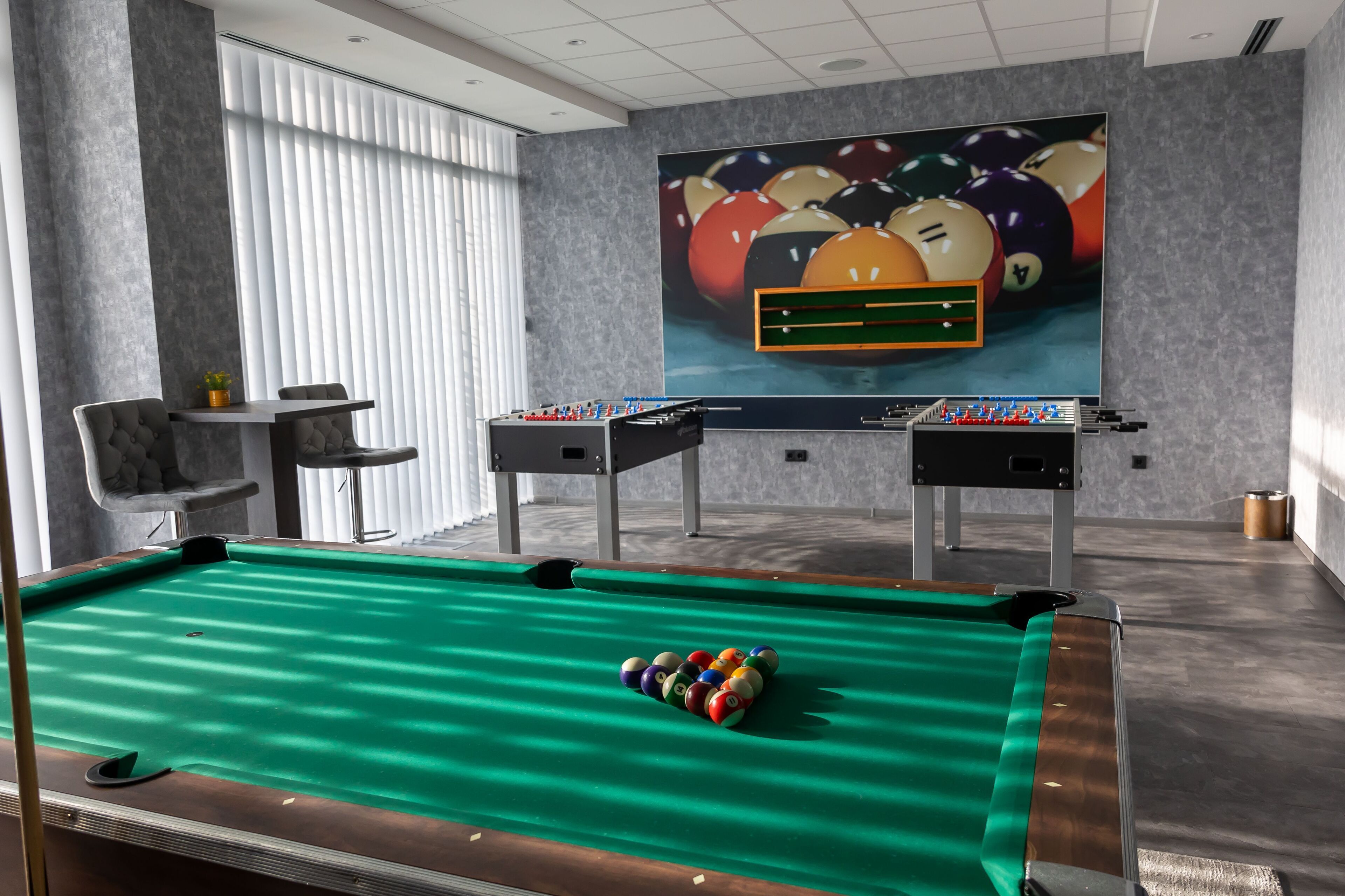 A pool table with a green felt surface and a cue ball, a foosball table, and a billiards table with a colorful ball arrangement.