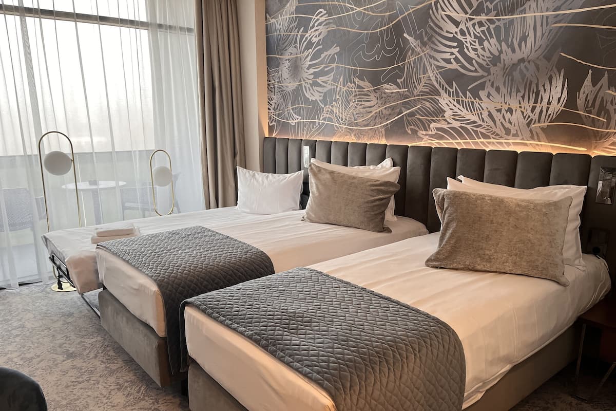 Habitación de hotel con dos camas, un ventanal grande con cortinas, un cabecero moderno y una obra de arte en la pared.