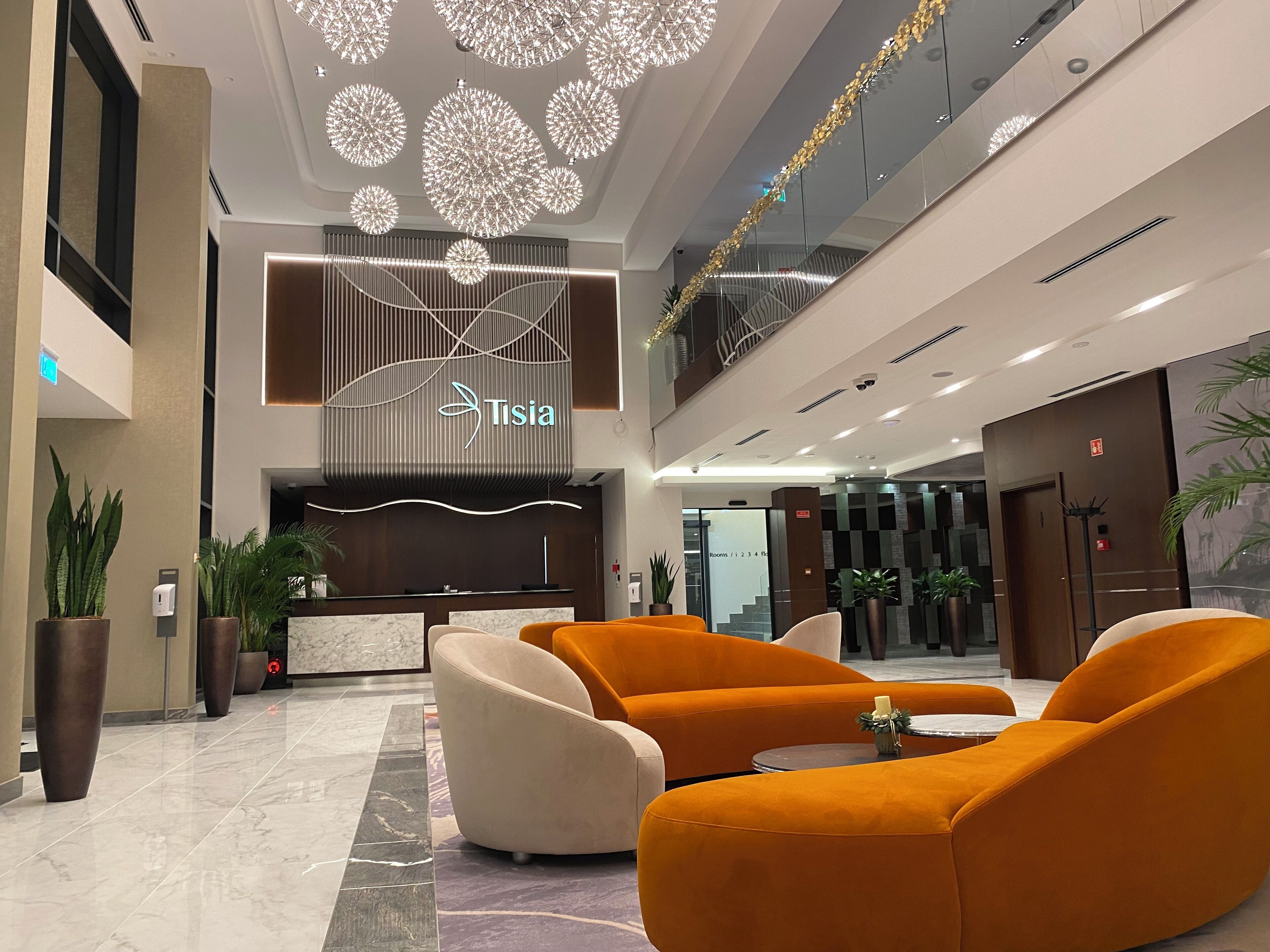 A modern hotel lobby with a reception desk, orange seating area, and a staircase leading to the upper floor.