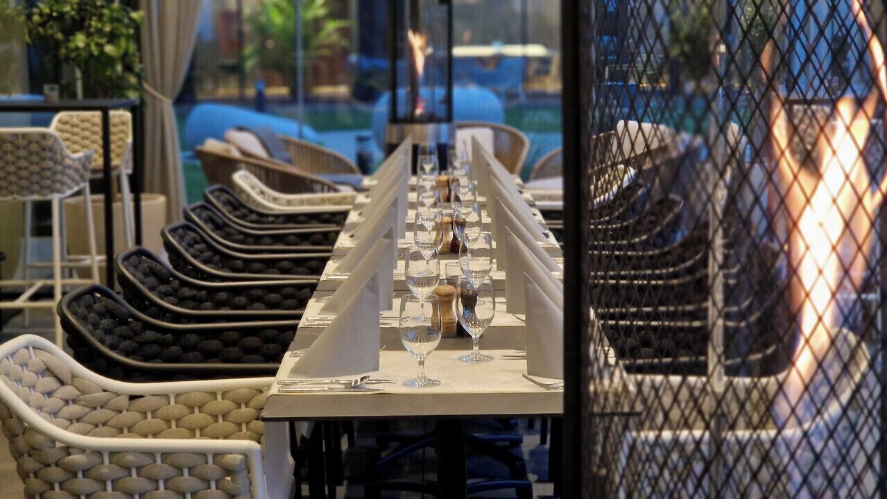 Mesa de jantar posta com copos de vinho e um menu, rodeada por cadeiras de vime.