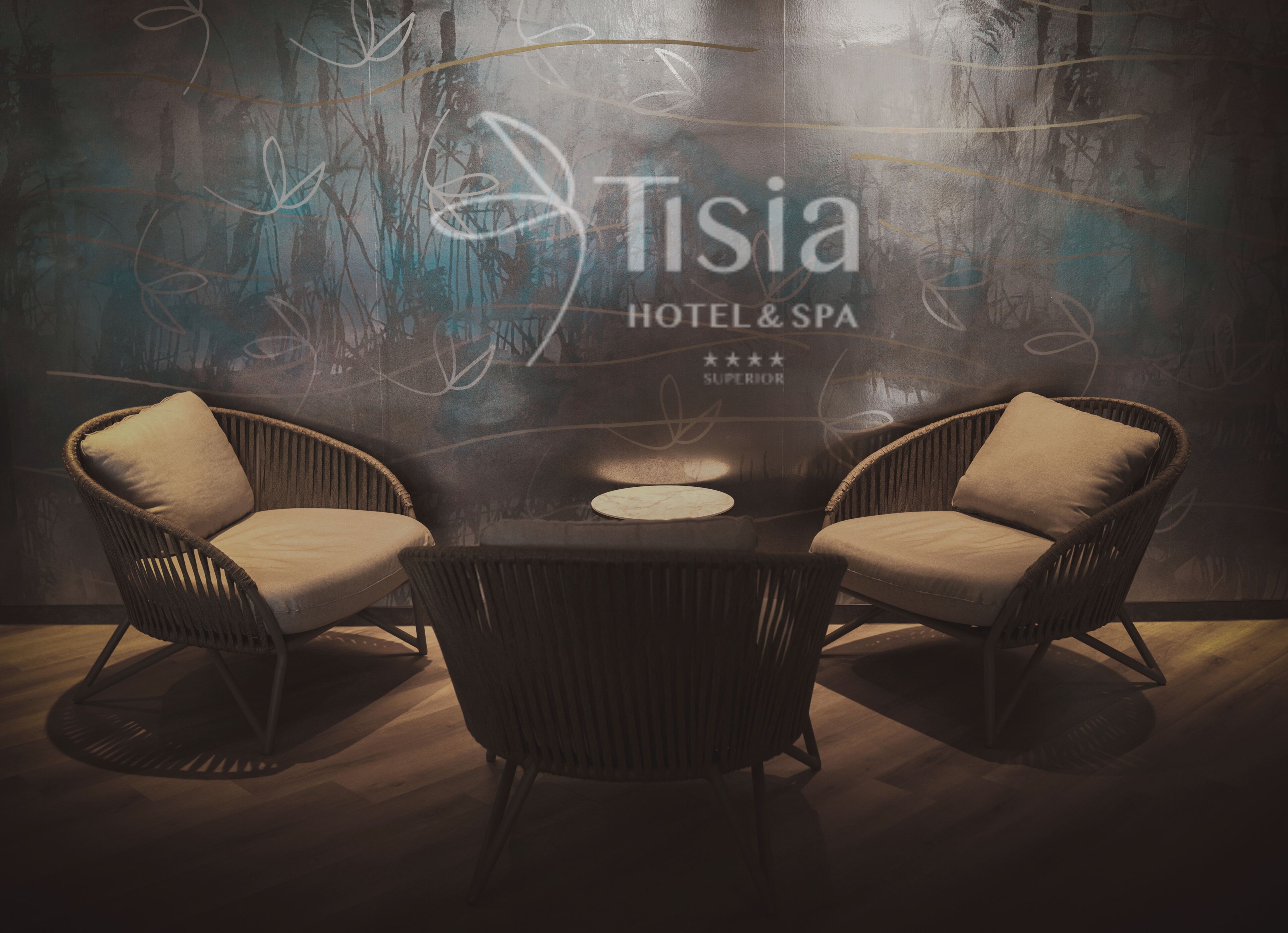 Een moderne hotelkamer met twee rieten stoelen, een ronde tafel en een muur met abstracte kunst en de hotelnaam 'Tisia'.