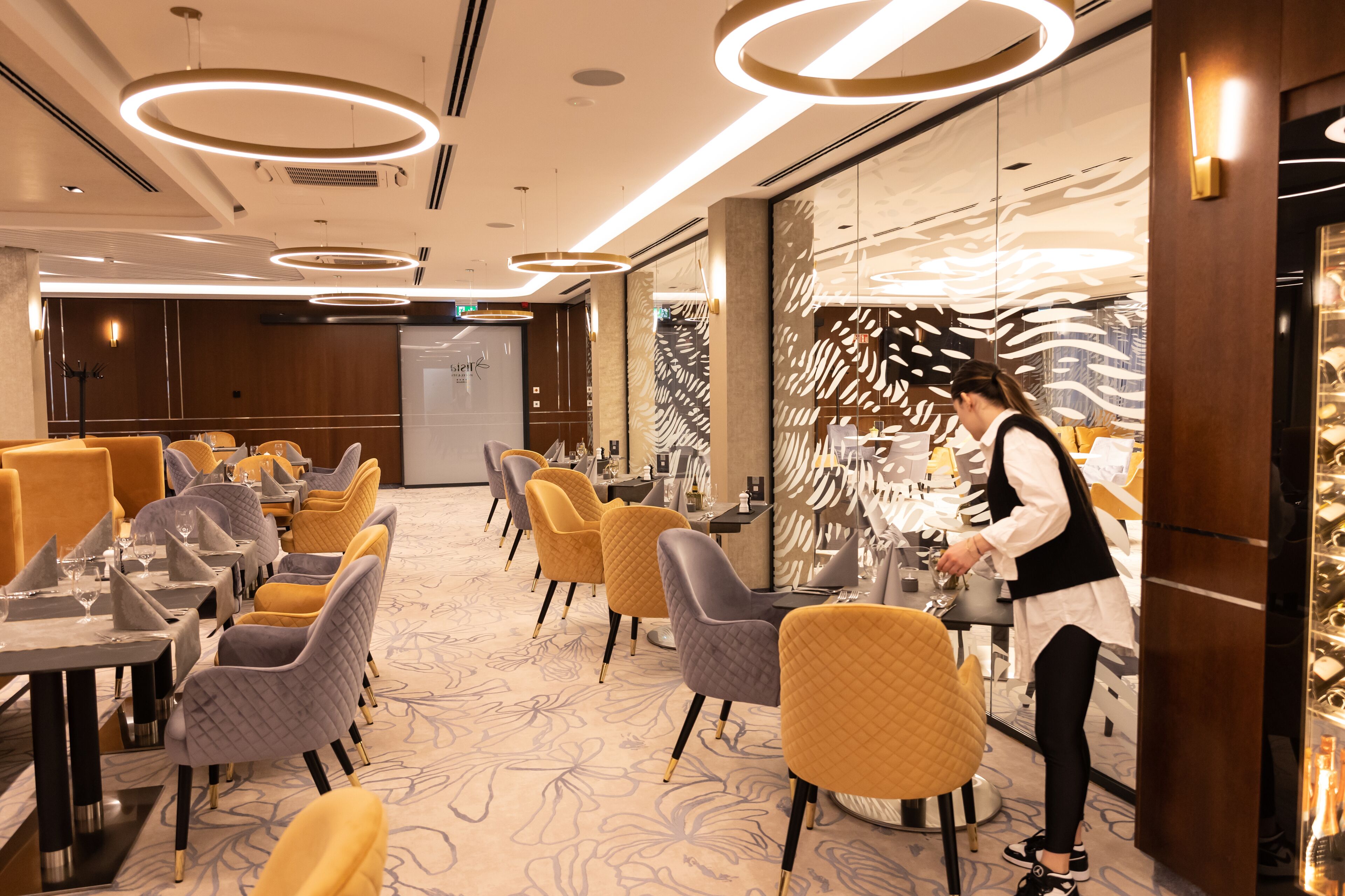 A modern dining area with round tables and chairs, a waiter attending to guests, and a decorative wall with a leaf pattern.