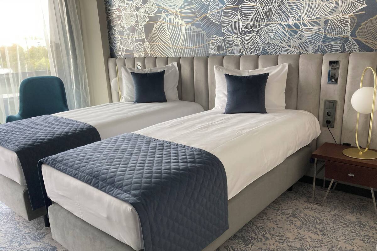Habitación de hotel con dos camas, una mesita de noche con lámpara y un papel tapiz estampado.