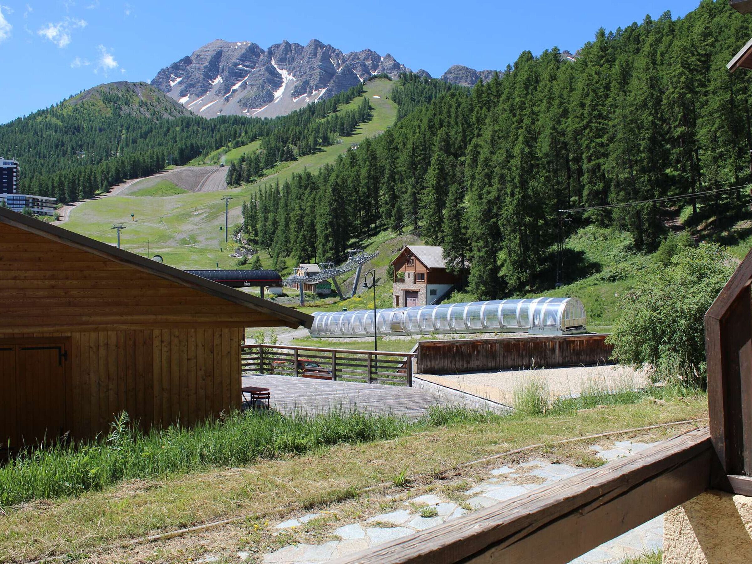 A wooden cabin with a view of a mountain range and a glass greenhouse structure.
