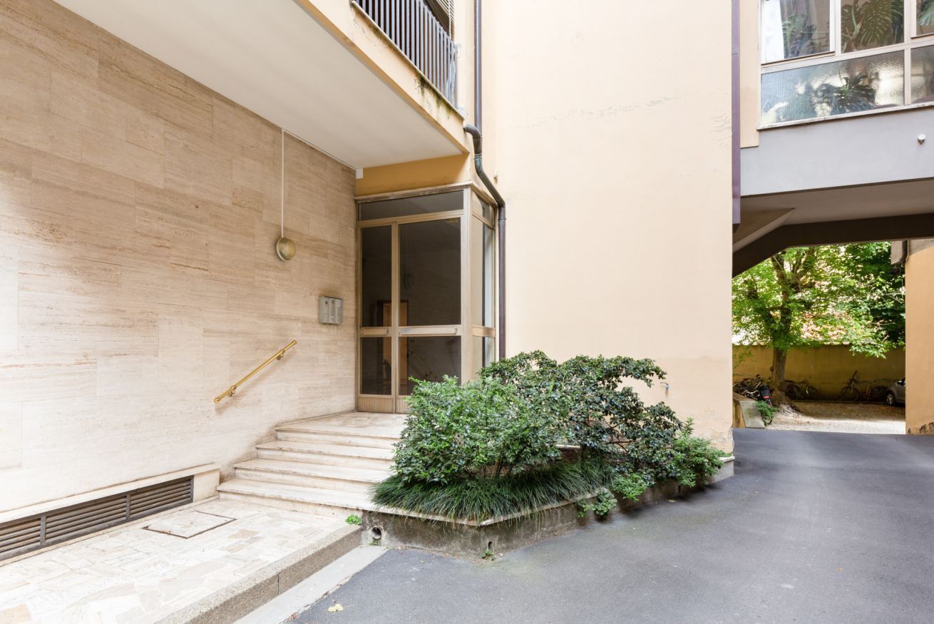 A building with a staircase leading to a door, a small garden area with bushes, and a paved ground.