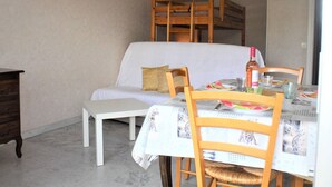 Un coin repas avec une table blanche dressée pour quatre personnes, des chaises en bois, un banc et, au fond, un lit superposé.