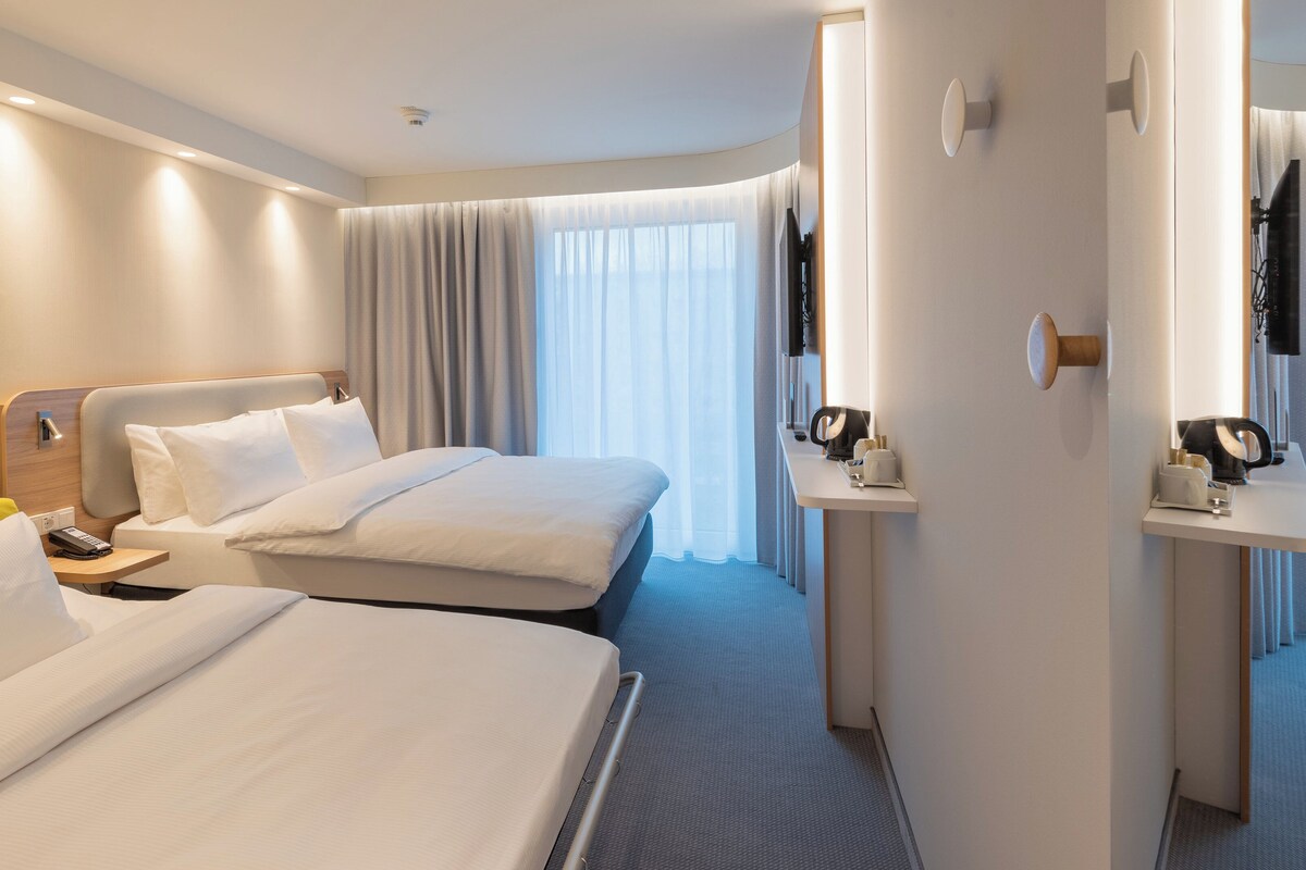 A hotel room with two beds, a bedside table with a phone, a small table with a coffee maker, and a large window with curtains.